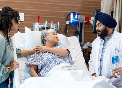 Older woman laying in a hospital bed holding her adult daughter's hand and speaking to a doctor