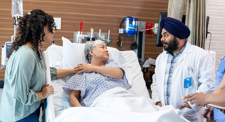Older woman laying in a hospital bed holding her adult daughter's hand and speaking to a doctor