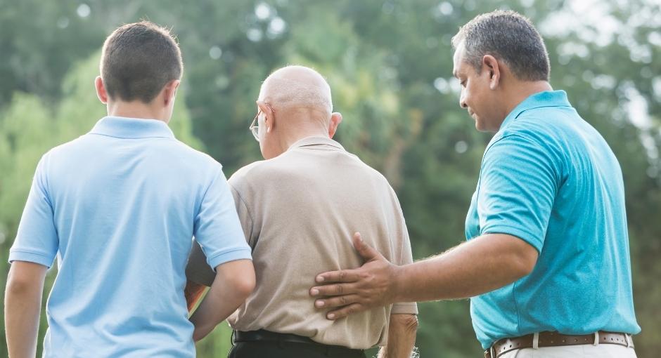 Multigenerational family of men with their backs to the camera. Grandfather stands in the middle with his grandson on the left and his son on the right. Son's left hand is placed on his father's back