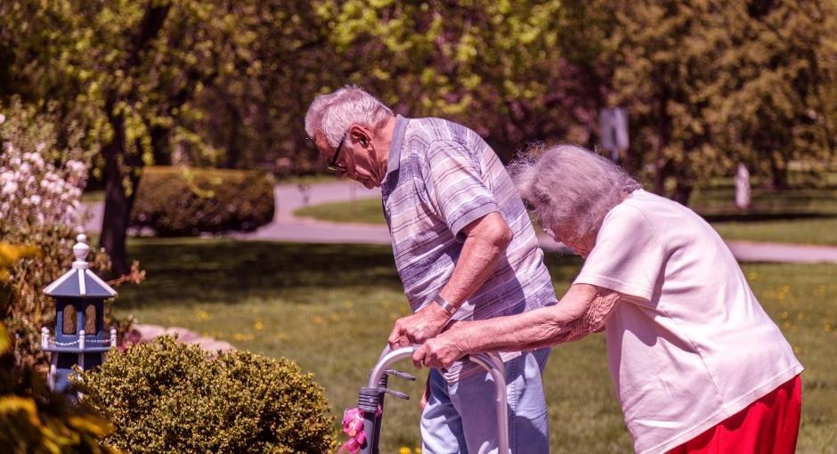 Older couple walking outside with assistance from walkers. Both of their backs are hunched over with stooped posture